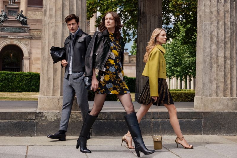 Three fashion models walk outdoors near stone columns, showcasing current apparel trends, including dresses, jackets, and boots.