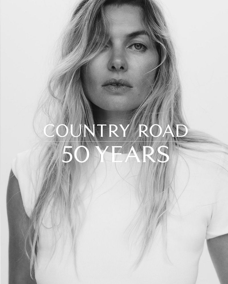 Black-and-white portrait of a blonde woman in a white t-shirt, featuring 'Country Road 50 Years' text overlay, with bright, soft lighting.