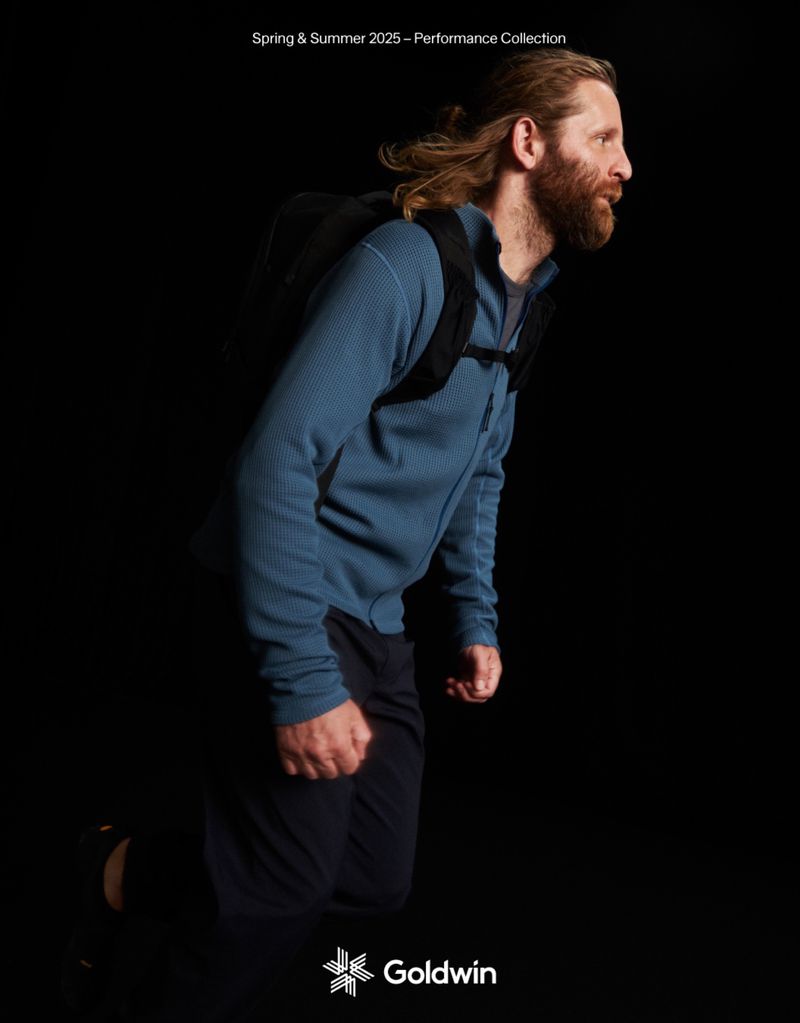 Man with backpack in motion. Model wears blue athletic jacket. Studio lighting highlights activewear for outdoor performance collection.