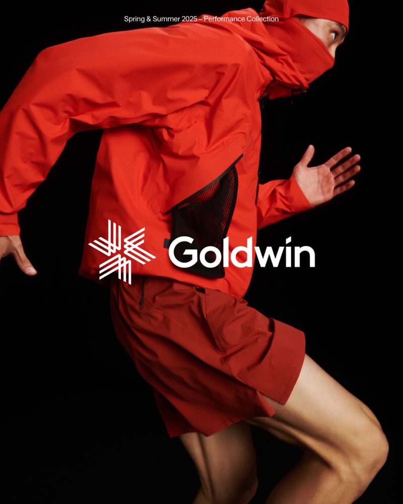 Person in motion wearing a red Goldwin hooded jacket and shorts against a black backdrop, showcasing athletic apparel.