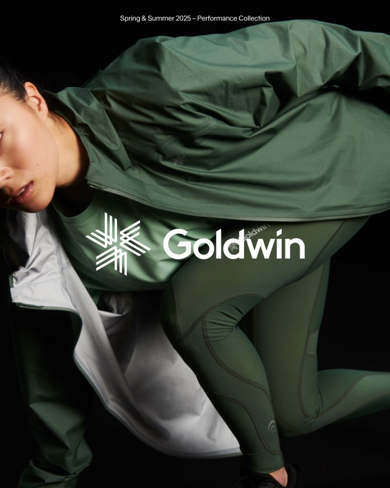 Fashionable athletic wear from Goldwin's Spring & Summer 2025 collection features a woman in a green jacket and leggings against a black backdrop.