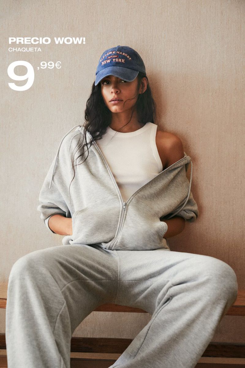 A young woman models a grey zip-up hoodie, sweatpants, white top, and blue baseball cap, seated indoors against a neutral backdrop.