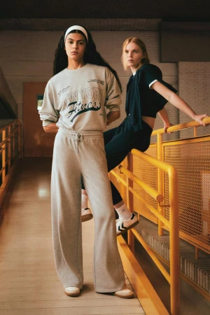 Two women in athletic wear pose indoors against a yellow metal railing and concrete walls. One woman wears a gray sweatshirt.
