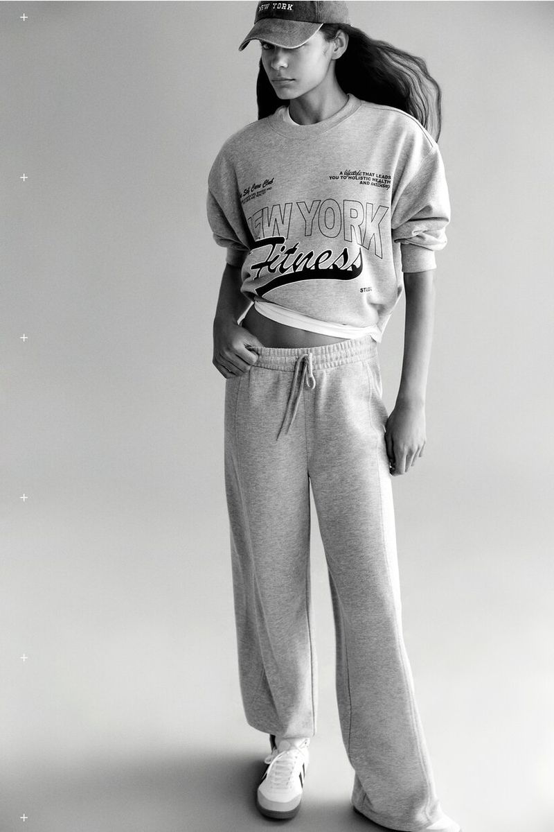 Young woman in a grey sweatshirt, baseball cap, and sweatpants posing in a studio with sneakers. Fashion and fitness apparel.