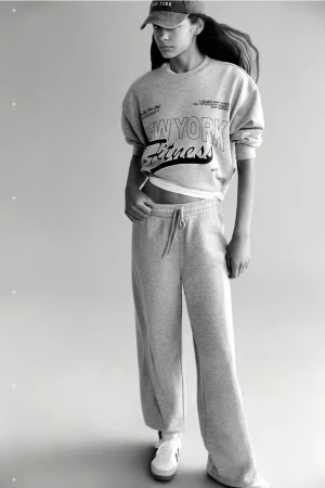Young woman in a grey sweatshirt, baseball cap, and sweatpants posing in a studio with sneakers. Fashion and fitness apparel.