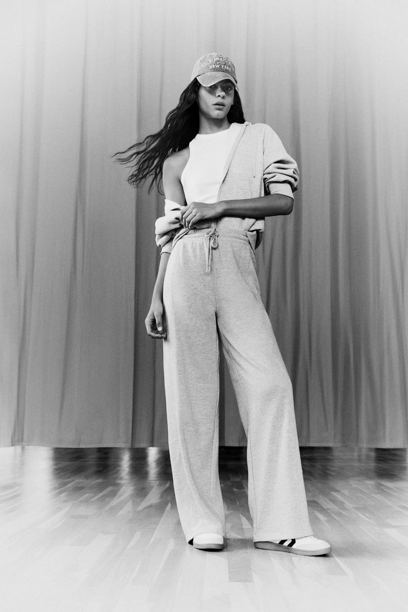Monochrome studio shot of a young woman in sportswear standing in front of a draped background, wearing a cap, tank top, and pants.