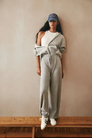 Model in grey tracksuit standing on a wooden bench against a neutral textured wall background