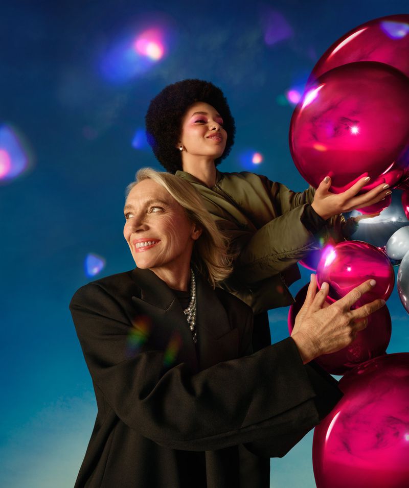 Two women pose with pink and silver balloons against a blue background with bokeh. The older woman wears a black blazer.