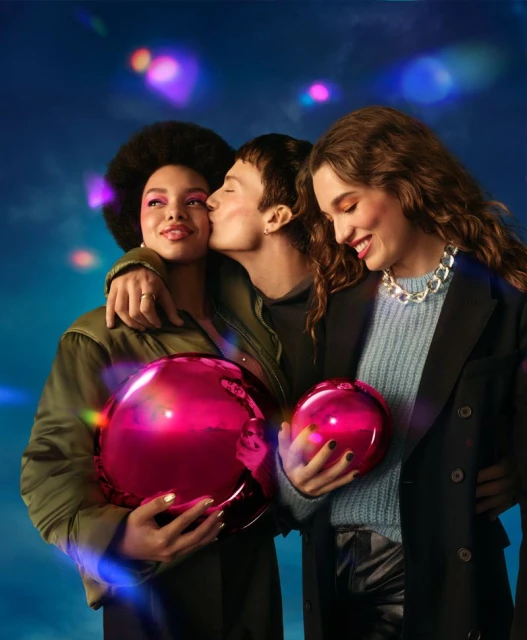 Three stylish adults pose indoors with vibrant pink orbs. The person in the middle kisses the cheek of the person on the left.