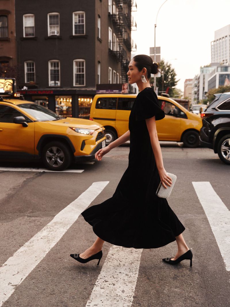 Elegant woman in black dress crossing city street in front of yellow taxis, holding clutch, wearing statement earrings and pumps.