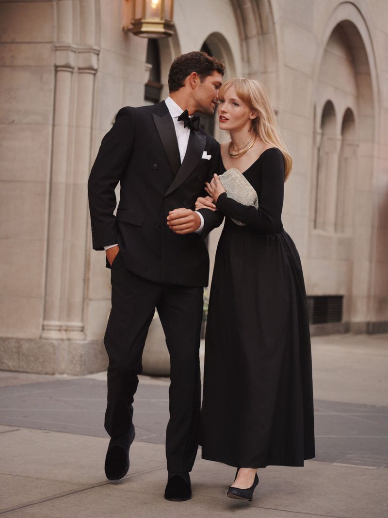 Man in a black tuxedo and woman in a long black evening dress walking together in front of an arched stone building.