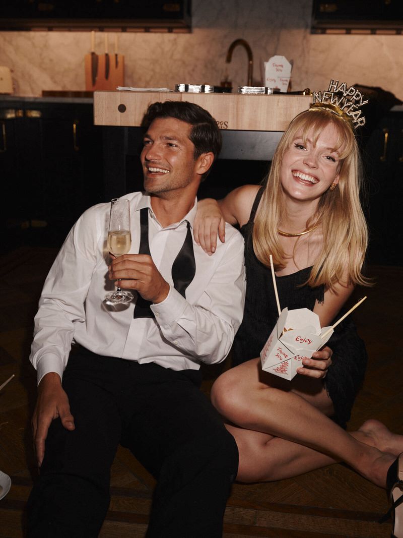 Smiling couple celebrates with champagne and takeout indoors. A man in a tux and a woman in a black dress pose casually on the floor.