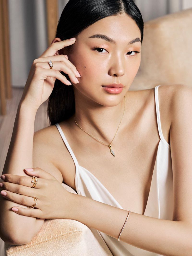 Portrait of an Asian woman with multiple gold rings, a gold necklace, and a bracelet while seated indoors against a neutral background.