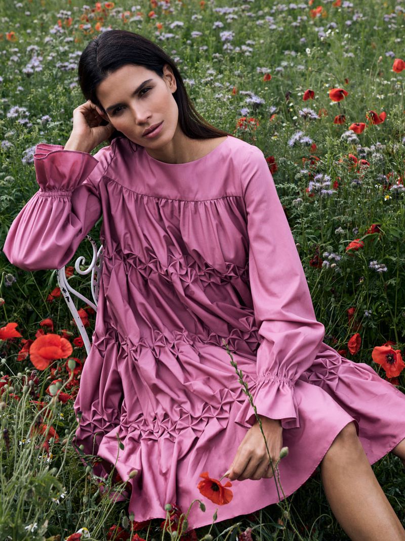 Adult female in a pink dress sits amongst wildflowers in a bright, natural setting, looking at the viewer with a relaxed expression.