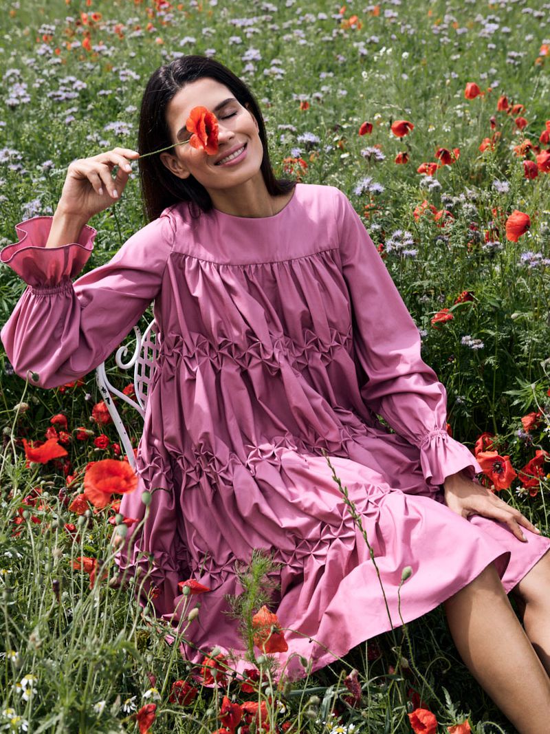 A woman in a pink dress sits in a field of wildflowers, holding a red poppy to her eye in natural light during daytime.