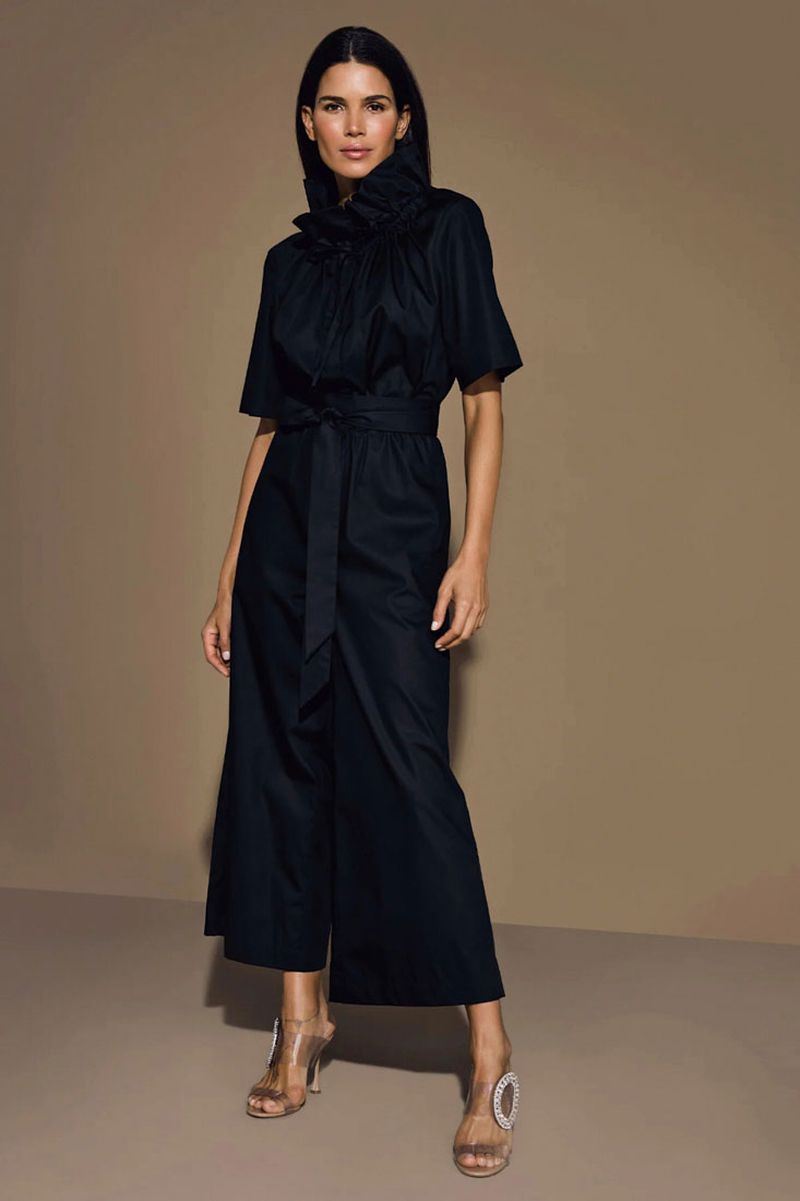 Full-length studio shot of a woman in a black jumpsuit with a tied waist and transparent heels, posing against a neutral background.