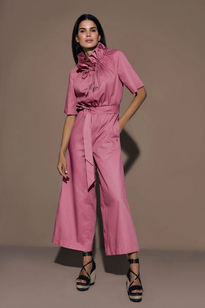 A full-length shot of a woman in a pink jumpsuit with ruffle details at the collar, a belt, and platform heels, against a beige backdrop.