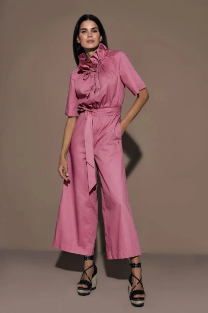A full-length shot of a woman in a pink jumpsuit with ruffle details at the collar, a belt, and platform heels, against a beige backdrop.