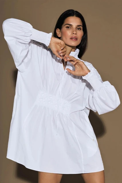 Woman in oversized white cotton shirt dress with smocked sleeves and a v-neck poses against a tan background in a studio shot.