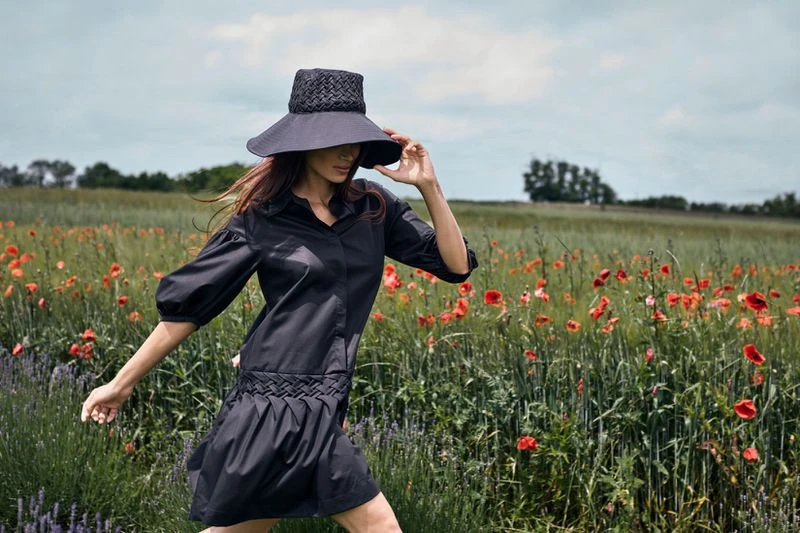 Woman in a black dress and sunhat walks through a field of red poppies under a cloudy sky, fashion and style.