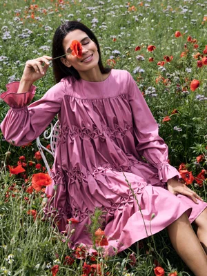 Raica Oliveira in a mauve gathered smock dress holding a red poppy flower over her eye while sitting in a wildflower meadow.