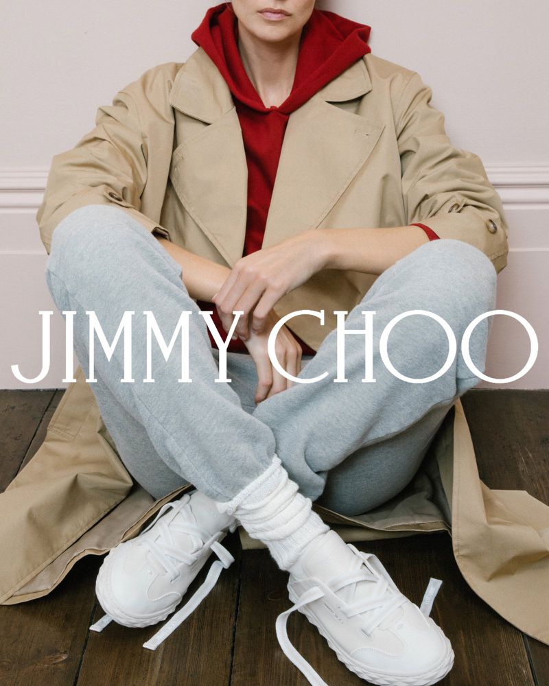 Jimmy Choo shoe advertisement featuring a person sitting cross-legged wearing white sneakers, grey sweatpants, and a trench coat.