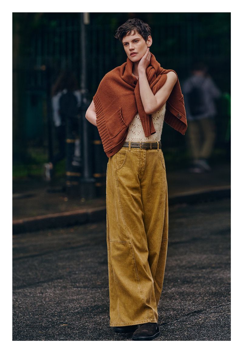 A young adult woman poses outdoors in a cream tank top, brown sweater, and wide-leg corduroy pants, with blurred figures in the background.