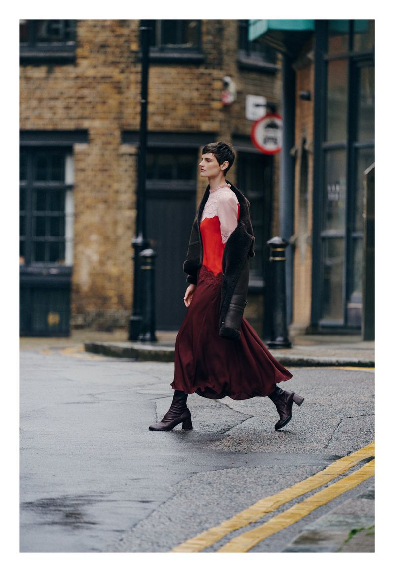 A woman in a red top and burgundy dress crossing a wet city street in a fashionable outfit with brick buildings in background.