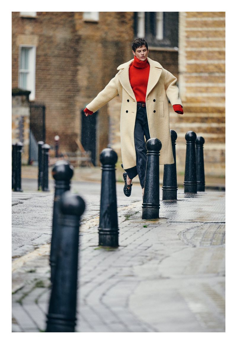 Stylish adult in an off-white coat, red sweater, and dark trousers balances on bollards in a wet urban setting with brick buildings.