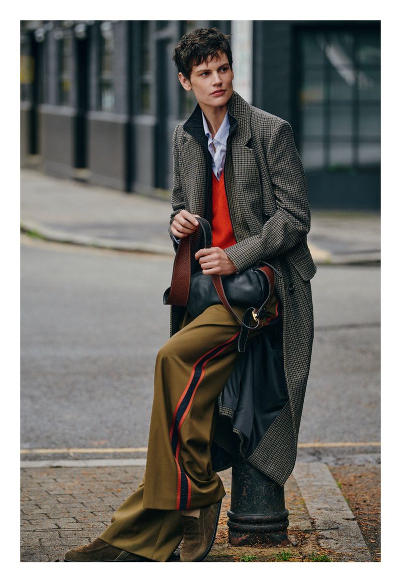 A stylish adult poses outdoors, wearing a long coat, earth-toned pants, and a sweater with a shoulder bag. The woman leans on a post.