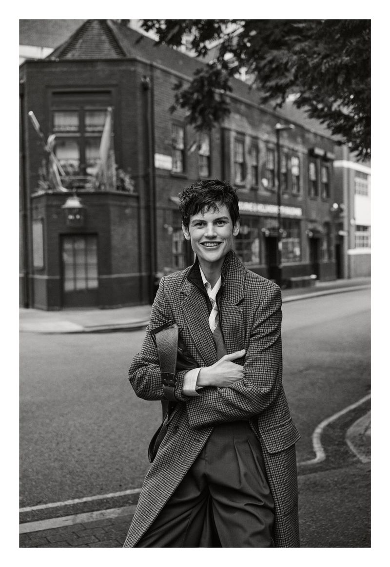 Monochrome full shot of smiling woman with short hair wearing a coat and carrying a bag, standing on a street with buildings.