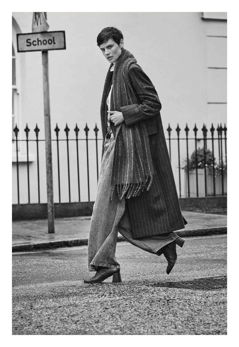 A woman walks across a crosswalk wearing an overcoat, scarf, and jeans in a monochrome outdoor fashion shot on a city street.