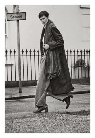 A woman walks across a crosswalk wearing an overcoat, scarf, and jeans in a monochrome outdoor fashion shot on a city street.