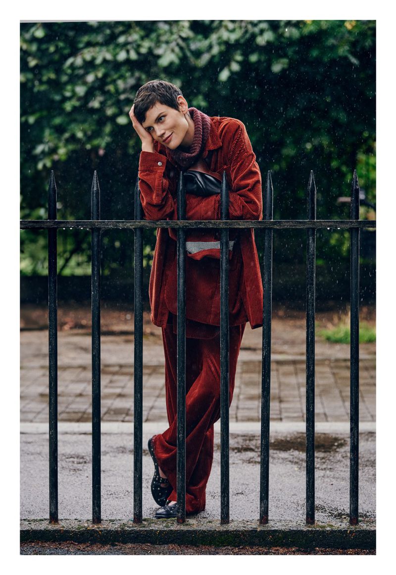 Woman in a burgundy outfit stands behind a black metal fence, resting her head on her hand with a soft smile. Rain is visible.