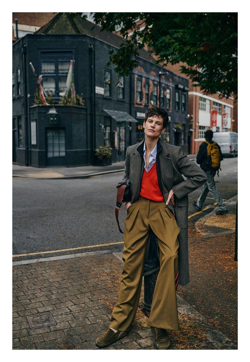 A woman poses in an urban outdoor setting, wearing olive-green trousers, a red top, and a tweed jacket, with a dark-colored building in the background.