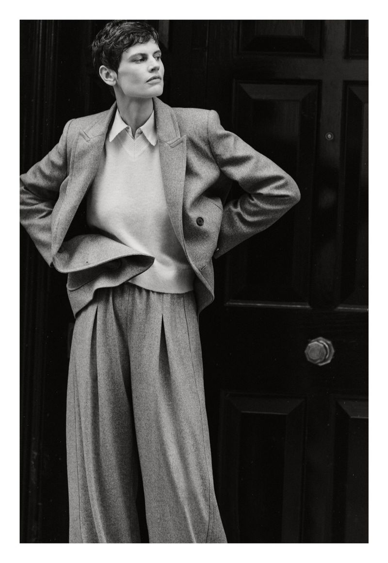 A woman in a grey suit stands against a dark door with her hands on her hips, posing for a fashion editorial in muted light.