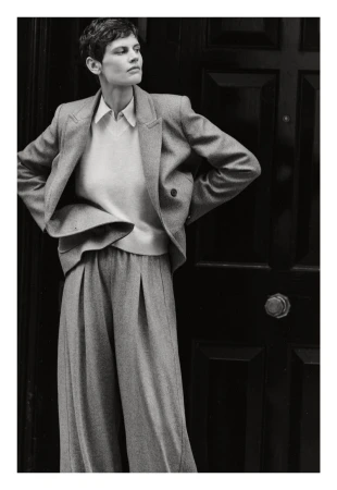 A woman in a grey suit stands against a dark door with her hands on her hips, posing for a fashion editorial in muted light.