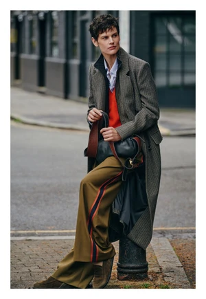 Saskia de Brauw wearing a long tweed coat and striped olive trousers perched on a street bollard for ME+EM.