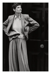 Model Saskia de Brauw wearing a tailored grey suit with wide-leg pleated trousers against a dark paneled door.