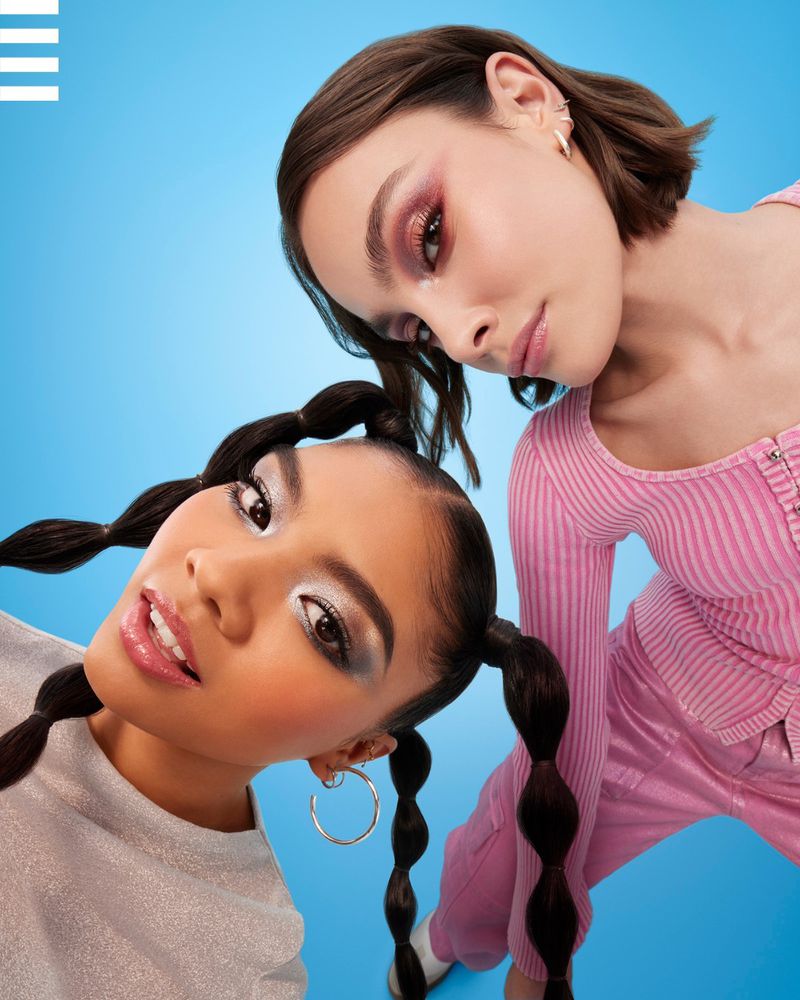 Two young women pose closely together against a bright blue background wearing stylish pink and silver eyeshadow makeup and retro-inspired outfits.