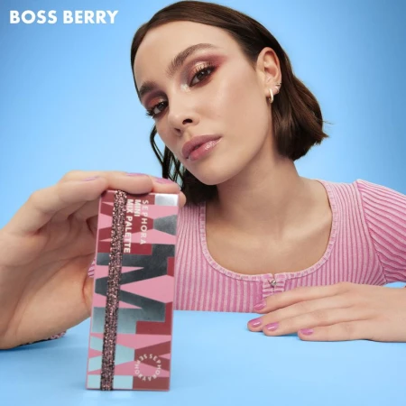 A woman models makeup while holding a Sephora makeup palette against a blue background. Berry hues are dominant in this beauty image.
