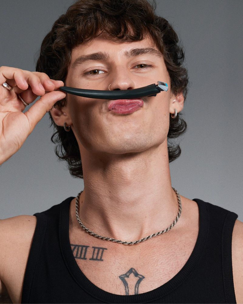 Man holds a styling wand to his face in a close up shot. He wears a tank top and silver chain necklace against a grey background.