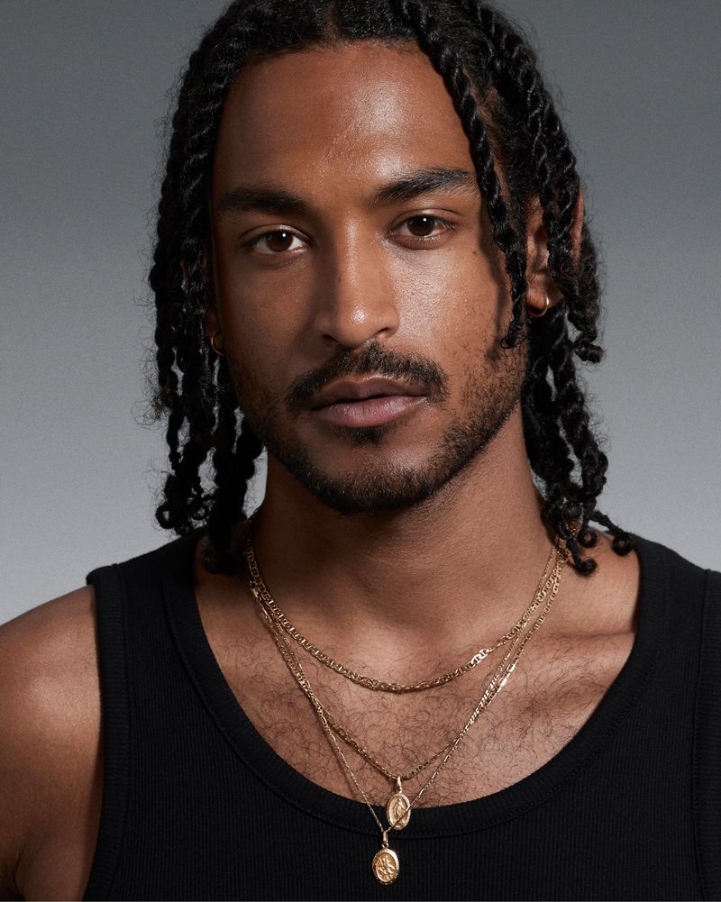 Close-up portrait of a Black adult male with dark braided hair, light beard, wearing a black top and gold necklaces against a grey background.