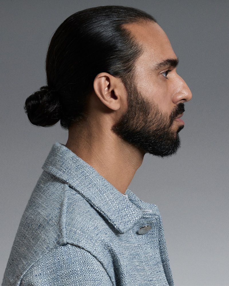 Side profile studio portrait of a bearded man with hair tied in a low bun wearing a light blue textured jacket against a grey background.