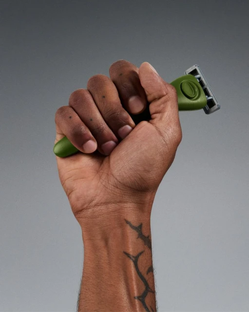 Close-up of a dark-skinned man's hand holding a green razor with a grey head against a light-grey studio backdrop, showcasing skin detail.