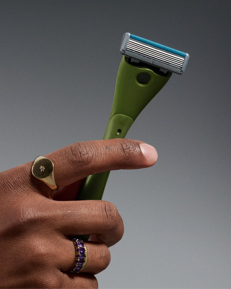 Close-up studio shot of a hand holding a green handled razor with a replaceable blade, adorned with gold rings against a gray backdrop.
