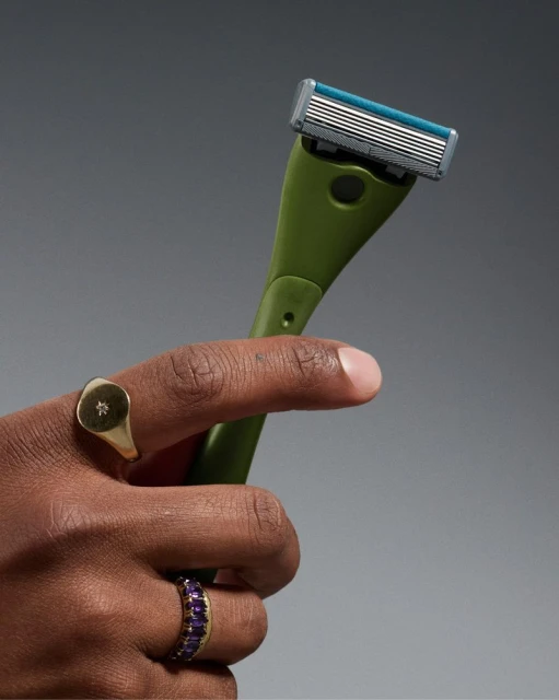 Close-up studio shot of a hand holding a green handled razor with a replaceable blade, adorned with gold rings against a gray backdrop.