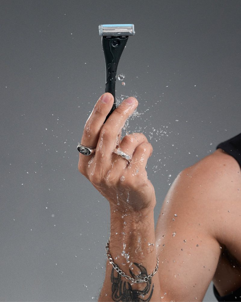 Razor in hand under running water, showcasing grooming and personal care against a grey backdrop, with a focus on male hygiene.