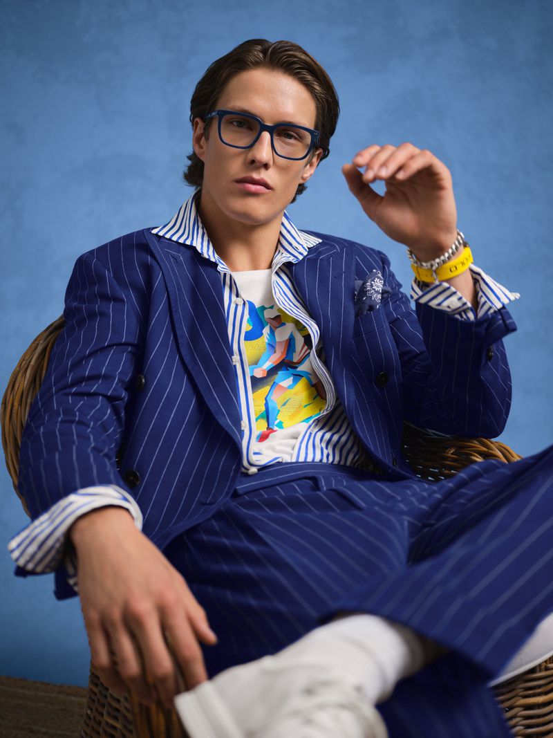 Gideon Yendell seated in a wicker armchair wearing a blue pinstripe suit, striped shirt, and eyeglasses against a blue backdrop.