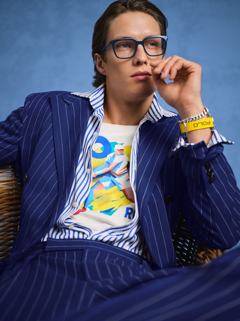 Adult male in a blue pinstripe suit and glasses sits in front of a blue backdrop. He wears a graphic tee and yellow bracelet.
